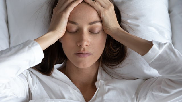 Frau liegt im Bett und hält sich mit beiden Händen den Kopf. Sie leidet unter Migräne. 